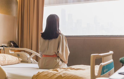 Back view of patient woman sitting on bed in hospital ward looking out at window