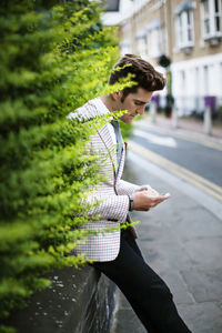 Businessman leaning on retaining wall and using smart phone on street