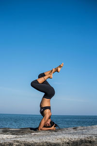 Young woman doing headstand at beach against sky