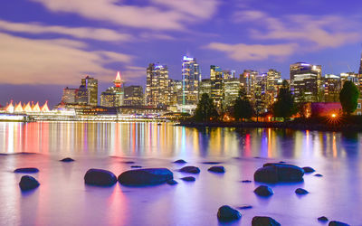 Illuminated buildings by river against sky at dusk