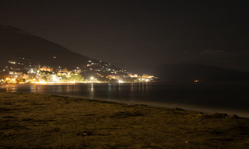 Illuminated city by sea against sky at night