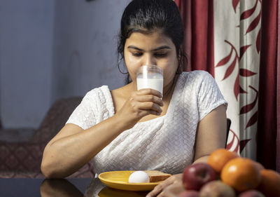 An indian asian woman drinking a glass of milk sitting beside a table in a domestic room