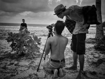 Full length of friends photographing man from camera at beach against cloudy sky