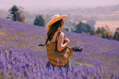 Woman with straw hat and tote standing in lavender flowers field