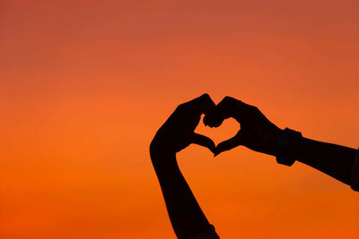 Silhouette hand holding heart shape against orange sky