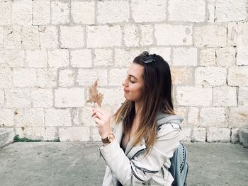 Teenage girl holding dry maple leaf while standing against wall