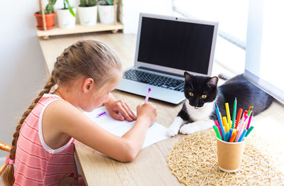 School girl doing homework at home, writing or drawing, cat lying nearby.