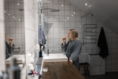 Side view of young blond woman brushing teeth while standing in bathroom