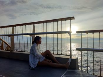 Woman sitting on railing against sky during sunset