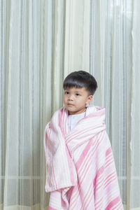 Boy wrapped in blanket standing against curtain at home