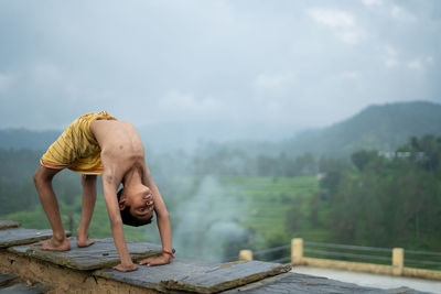 Full length of shirtless man with arms outstretched against sky