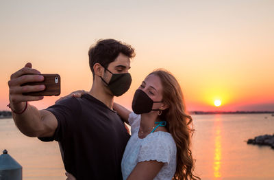 Young woman photographing while standing on shore against sky during sunset