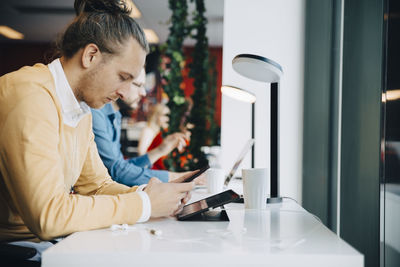 Side view of mid adult businessman using smart phone while sitting at desk in office