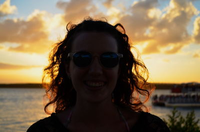 Close-up of smiling young woman wearing sunglasses at sea against sky during sunset