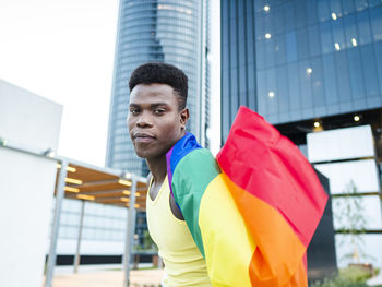 Young man wrapped in rainbow flag by modern buildings