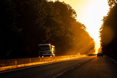 Cars on road by trees against sky during sunset