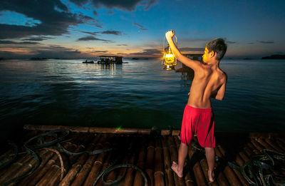 Rear view of shirtless boy holding oil lamp on wooden raft in sea at night