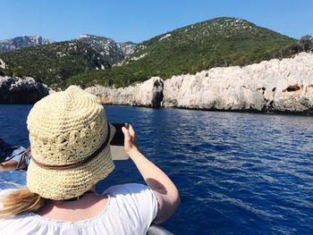 Rear view of woman photographing mountains from boat in sea