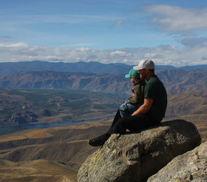 Full length side view of father and son sitting on rock at mountain peak