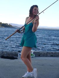 Smiling young woman holding fishing rod by sea against sky