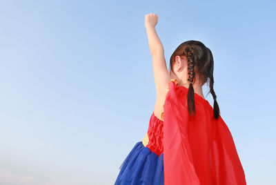 Low angle view of girl with hand raised standing against clear blue sky