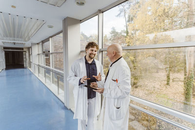 Smiling male doctor discussing with senior colleague standing in corridor at hospital