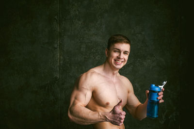 Portrait of shirtless man with bottle gesturing thumbs up against wall