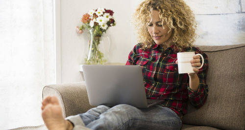 Smiling woman using laptop while holding coffee cup at home