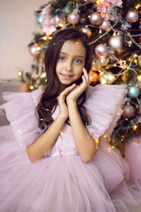 Beautiful baby girl with big eyes in a pink dress is sitting at the christmas tree at home