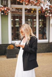 Young fashionable blonde woman in a black suit using mobile and walking through shops of the city