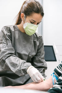 Professional female medical specialist in protective uniform and mask preparing arm of anonymous patient for blood test in modern lab