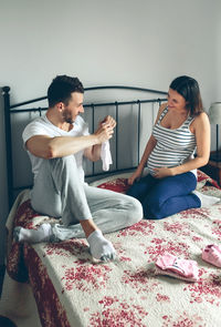 Man with pregnant women looking baby clothing while sitting on bed at home