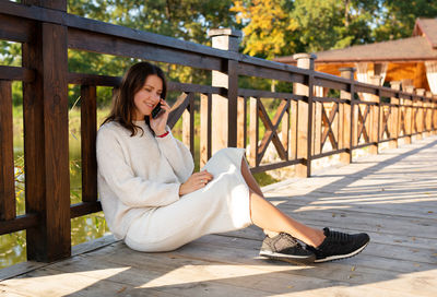 The girl is talking on the phone while sitting on the street on a wooden bridge