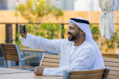 Man wearing traditional clothing taking selfie at outdoor cafe