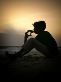 Teenage boy sitting at beach against sky during sunset