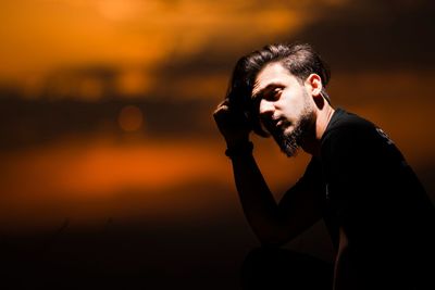 Young man looking away while sitting against sky during sunset