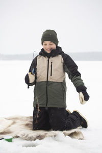 Happy boy in warm clothing kneeling while fishing on frozen lake