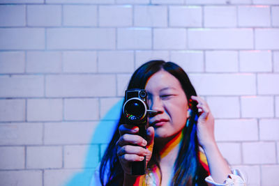 Young woman looking through video camera filming in front of wall