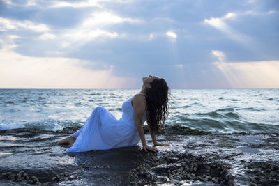 Side view of young woman sitting at beach against cloudy sky