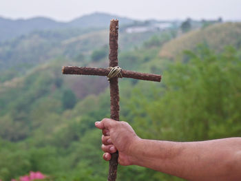 Cropped hand holding wooden cross against mountain