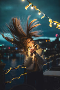 Portrait of smiling young woman tossing hair while holding illuminated lighting equipment at night