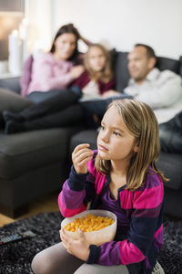 Girl having snacks while watching tv on floor with family in background