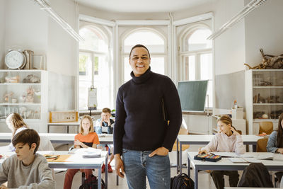 Portrait of happy male teacher with hand in pocket standing amidst students in classroom