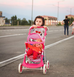 Cute girl pushing doll in baby stroller on sidewalk against sky