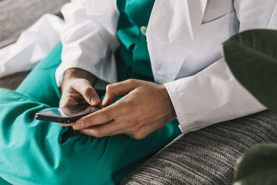 High angle of crop faceless male general practitioner in uniform sitting on couch in hospital and browsing mobile phone