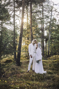 Full length of newlywed couple standing by trees at forest during wedding