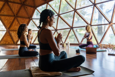 People in front of her teacher in sportswear while are seating in lotus pose with namaste hands while practicing breathing exercise during yoga session in studio