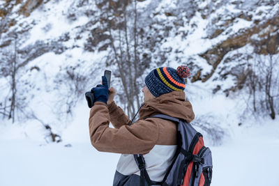 Young man  with backpack walking in snow among rocks in winter taking photos with smartphone hiking