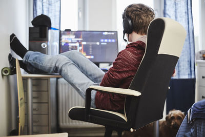 Teenage boy sitting on chair with feet up by computer while listening music through headphones at home