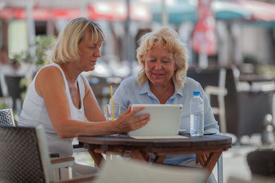 Mature woman showing digital tablet to friend at sidewalk cafe